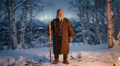 Anciano ainu con barba blanca y traje tradicional attus bordado con patrones geométricos morew, sosteniendo bastón ritual en bosque de abedules nevado de Hokkaido, luz suave invernal