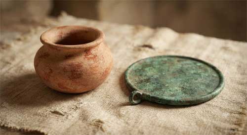 Pequeño recipiente de cerámica antigua con restos de pigmento rojo y espejo de bronce patinado dispuestos sobre tela de lino envejecida, luz suave lateral creando sombras delicadas, atmósfera de reverencia silenciosa hacia objetos personales del pasado