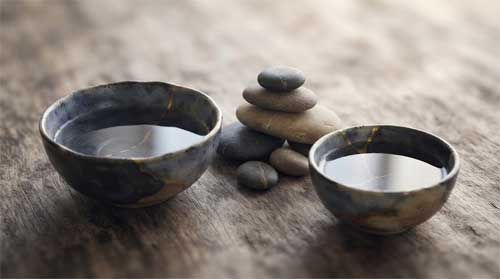 Composición minimalista de dos cuencos de cerámica rústica con agua reflejando luz suave, piedras lisas dispuestas en equilibrio, atmósfera serena de concordia
