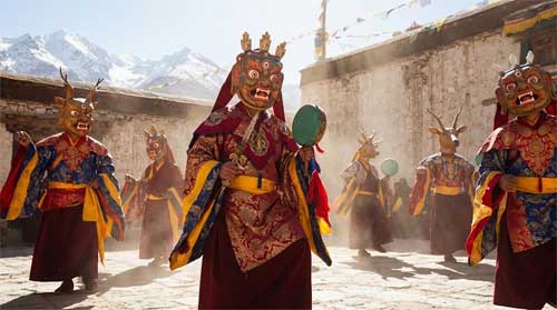 Monjes tibetanos realizando danza Cham con máscaras coloridas de deidades iracundas, vestidos con sedas bordadas, en patio de monasterio bajo cielo azul intenso