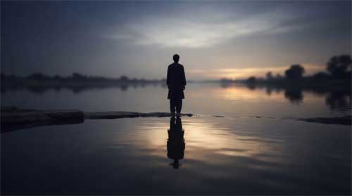 Silueta reflejada en superficie de agua tranquila al atardecer, luz suave creando distorsión sutil, atmósfera de introspección y mirada externa