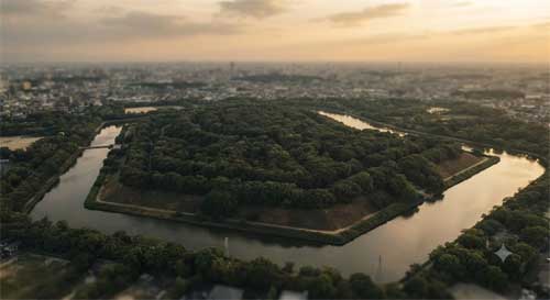 Vista aérea del kofun en forma de ojo de cerradura rodeado de triple foso y bosque denso, luz suave de atardecer resaltando geometría sagrada y escala monumental, atmósfera de misterio y poder ancestral sellado bajo vegetación
