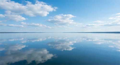 Superficie de agua completamente plana reflejando el cielo azul y nubes blancas con nitidez perfecta, sin ondas ni distorsiones, luz natural brillante, sensación de calma absoluta y claridad