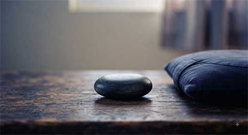 Piedra oscura equilibrada sobre madera junto a zafu simbolizando postura como presencia iluminada