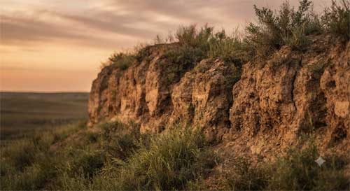Tramo antiguo de muralla Qin de tierra apisonada erosionada por tiempo, con vegetación creciendo entre grietas, luz dorada de atardecer resaltando textura orgánica y escala humana de construcción, atmósfera de obra colectiva anónima más que monumento imperial