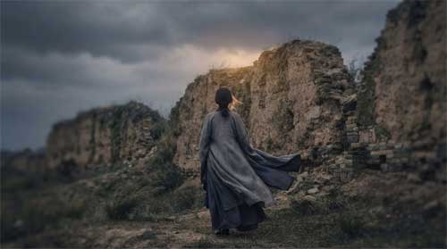Silueta femenina solitaria ante muralla antigua bajo cielo nublado, viento moviendo vestimenta tradicional, atmósfera de duelo profundo y resistencia silenciosa