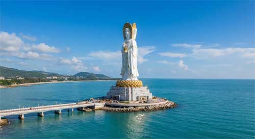 La monumental Estatua de Guanyin de Nanshan emergiendo del mar azul turquesa bajo un cielo despejado, con el templo en la costa al fondo