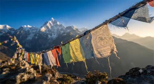 Banderas de oración tibetanas ondeando contra cumbres nevadas del Himalaya bajo cielo azul intenso, luz dorada de amanecer resaltando colores desvaídos por viento, atmósfera de sacralidad y resistencia silenciosa