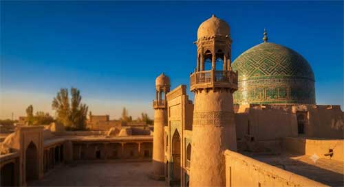 Arquitectura islámica tradicional de Kashgar con minaretes de adobe y cúpulas verdes bajo cielo azul intenso, luz cálida de atardecer resaltando texturas terrosas, atmósfera de herencia centroasiática en suelo chino