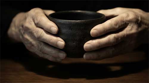 Detalle de manos ancianas sosteniendo cuenco de té Bizen oscuro sin esmalte sobre superficie madera oscura pulida, iluminación tenue lateral resaltando textura áspera cerámica y arrugas piel, atmósfera silenciosa y reverente