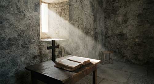 Vista interior de una celda monástica sencilla con luz entrando por una ventana estrecha, iluminando una cruz de madera y un libro abierto