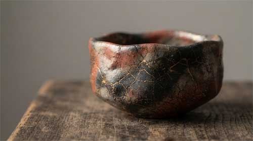 Cuenco raku handmade asimétrico con esmalte craquelado tonos óxido y negro mate, colocado sobre loseta madera antigua desgastada, luz lateral suave revelando irregularidades superficie y textura táctil, fondo neutro gris piedra