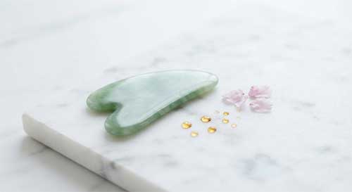 Herramienta de Gua Sha de jade verde suave descansando sobre una superficie de mármol blanco, acompañada de gotas de aceite esencial y pétalos de flor