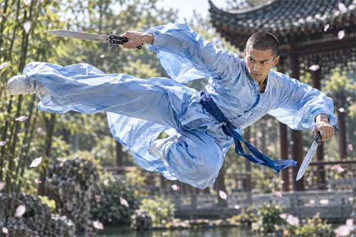 Practicante de kung fu en pleno salto acrobático sosteniendo dos machetes mariposa, con la tela de su uniforme ondeando como alas