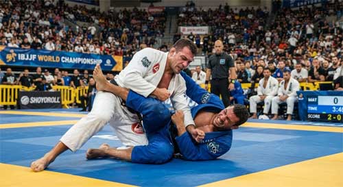 Dos atletas de BJJ con kimonos blancos y azules luchando intensamente sobre un tatami, rodeados de una multitud animada en un gran pabellón deportivo