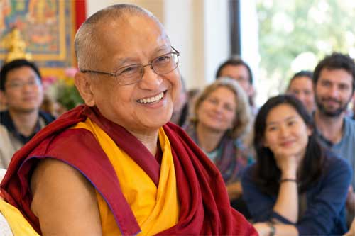 Lama Zopa Rinpoche sonriendo ampliamente con sus características gafas, rodeado de estudiantes diversos en un entorno monástico occidental