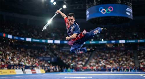 Atleta de Wushu en pleno salto acrobático con una espada Jian, con los anillos olímpicos proyectados sutilmente en el fondo de un estadio moderno